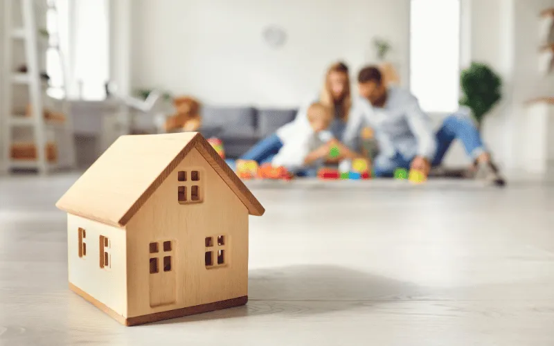 Wooden toy house in foreground with family playing together in bright living room