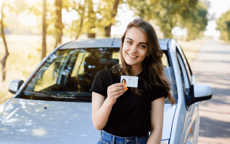 Young woman proudly showing her driver’s license in front of a car