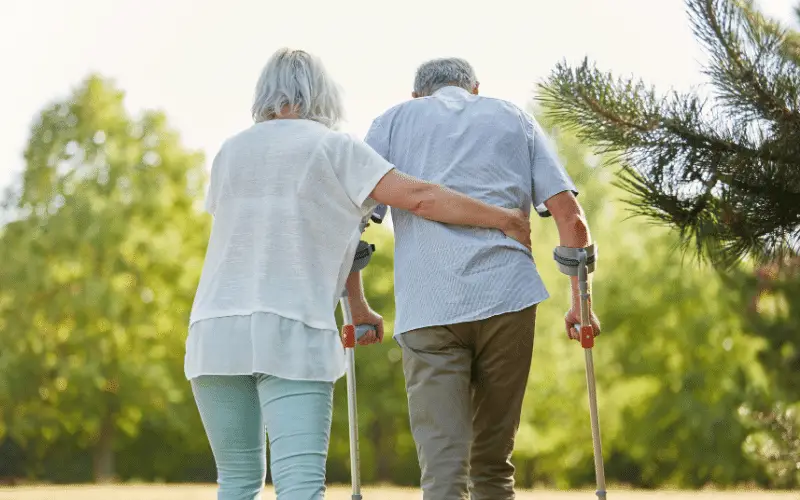 Senior woman supporting elderly man with crutches while walking outdoors