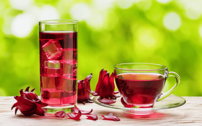 Glass of iced hibiscus tea and cup of hot hibiscus tea with petals on wooden table