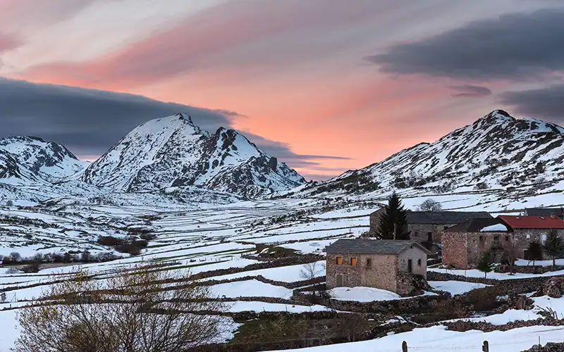 Idyllic snow-covered mountain village in northern Spain at sunset, with rugged peaks, scattered stone houses and pastel winter sky