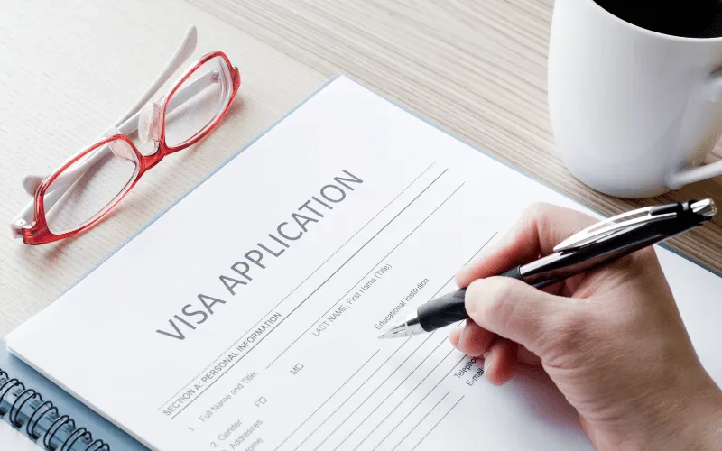 Hand filling out visa application form at desk with coffee and glasses nearby