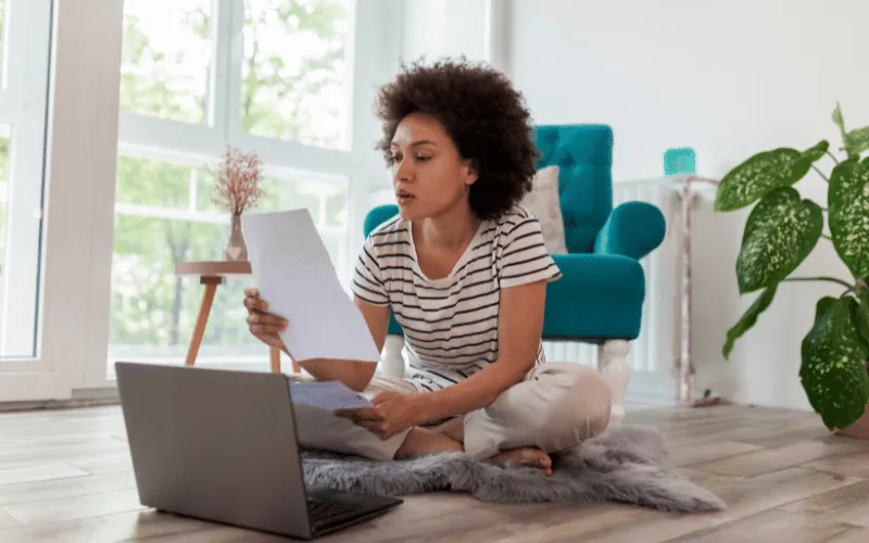 Young woman reviewing bills at home with laptop on the floor