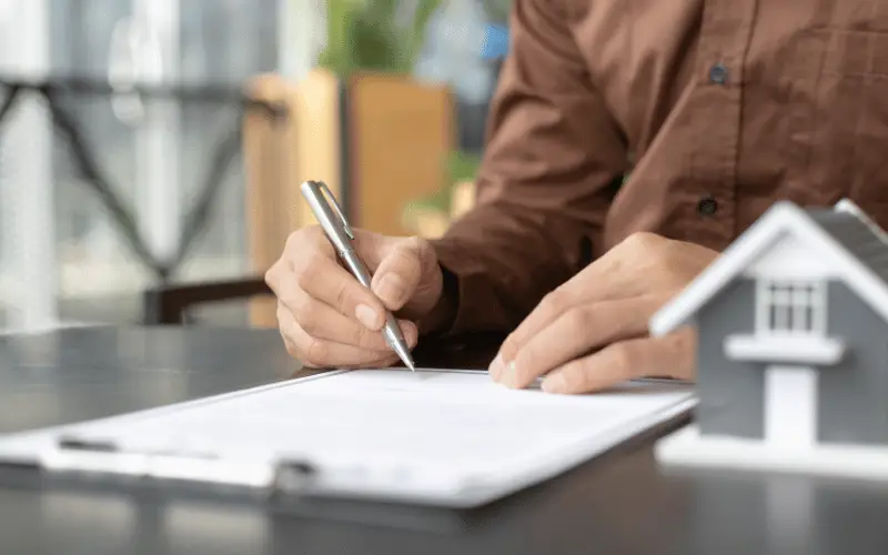 Man signing real estate contract at desk with house model for property purchase, rental agreement, or mortgage deal