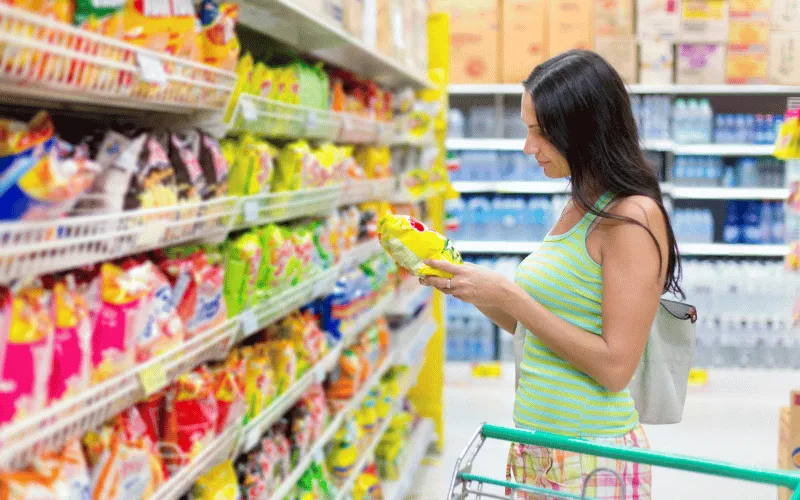 Shopper reading snack label in supermarket aisle filled with colorful packaged foods