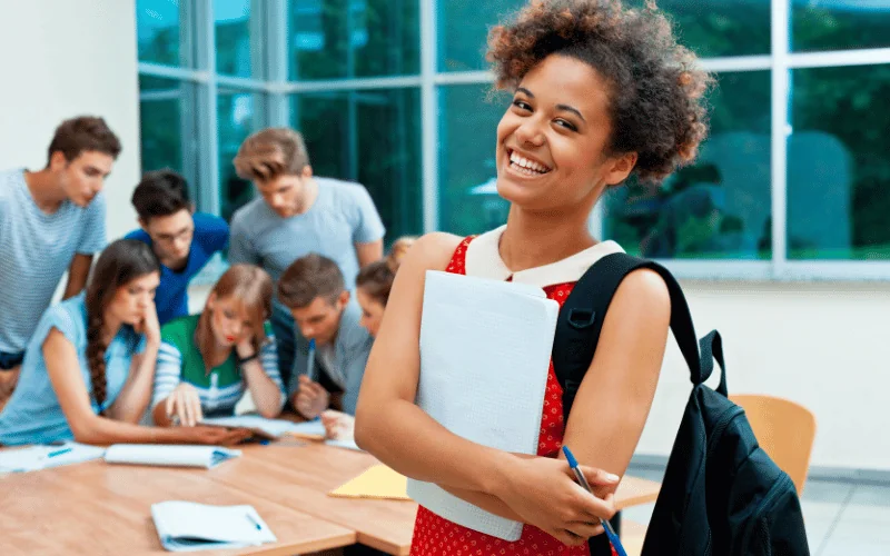 Smiling university student with backpack in classroom with group studying in background