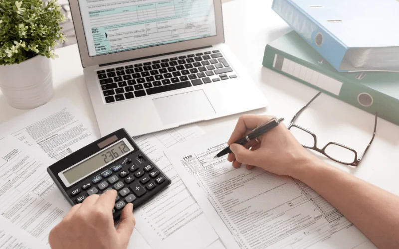 Person calculating taxes at home with documents, calculator, and laptop on desk