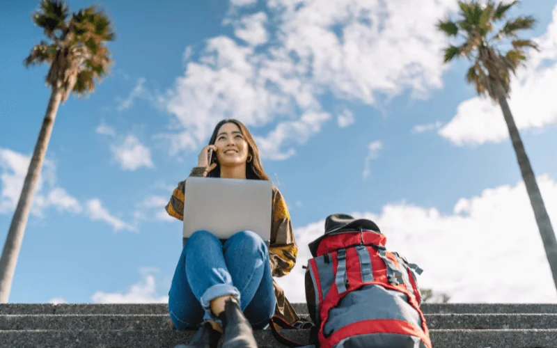 Young digital nomad working on laptop outdoors with backpack and palm trees